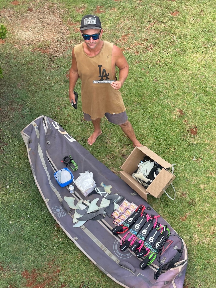 Overhead view of surfboard carry bag, and a bunch of gear layed out on it with a man standing there on the lawn in tank top