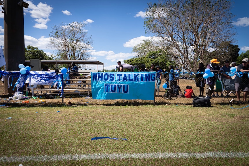 A blue banner which says 'whos taking tuyu' on a fence among blue balloons on a fence