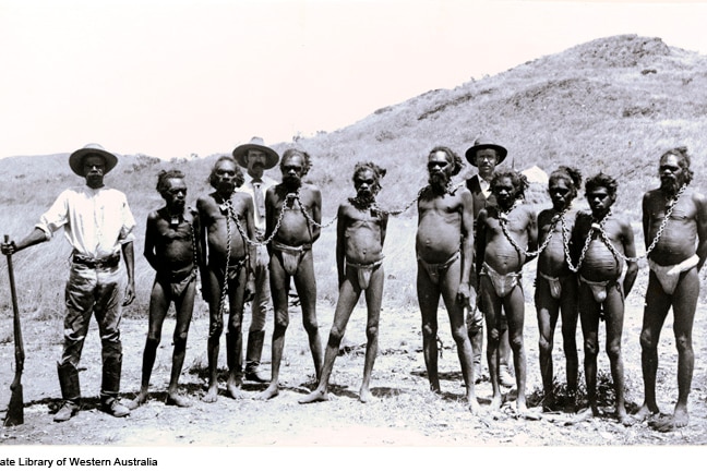 A group of chained Indigenous men in lion cloth, standing with Indigenous man wearing trousers, shirt, hat, holding gun.