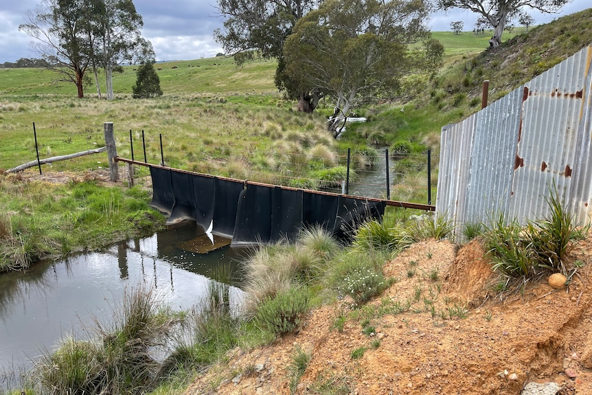 A farm fence built using old conveyor belt, roofing iron and big metal supports crosses a creek.