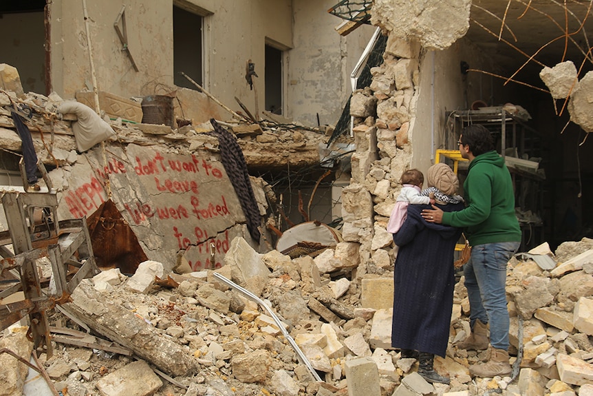 A couple with young baby daughter stand in destroyed building and look at red graffiti message.