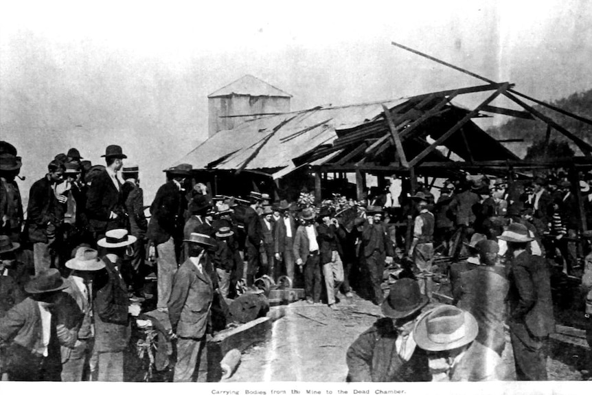 A large group of men look on as other men carry a stretcher with a broken shed in the background.