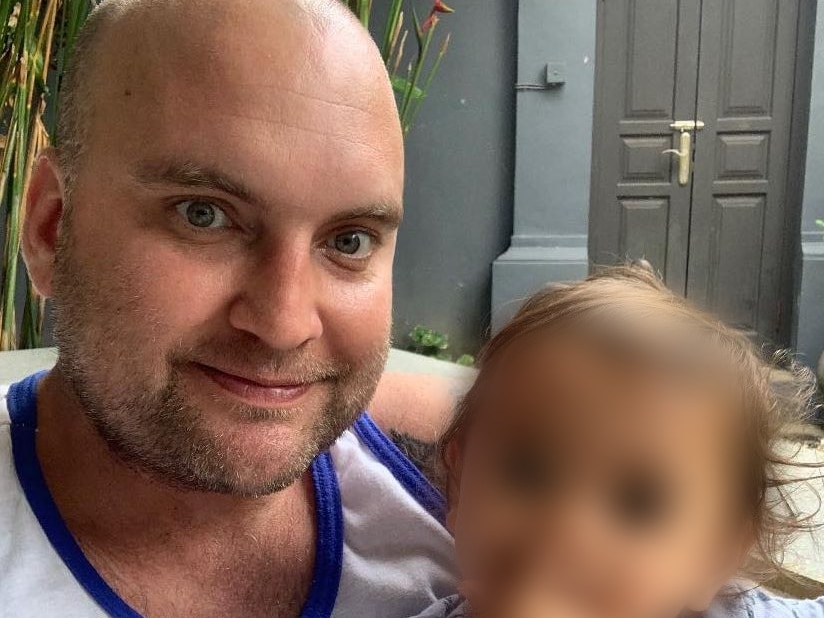 A father wears a singlet and holds his baby who has his face blurred