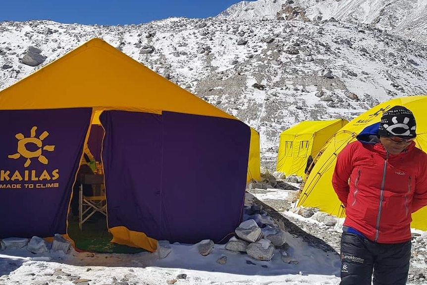 A man standing in front of tents and snow capped mountains