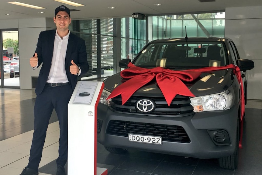 Selby Lee-Steere stands in front of a car with a giant red ribbon on its hood in a showroom.