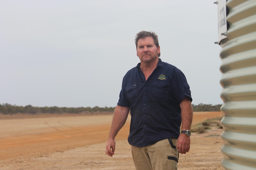 He stands on the red dirt airstrip near a water tank, scowling at the camera