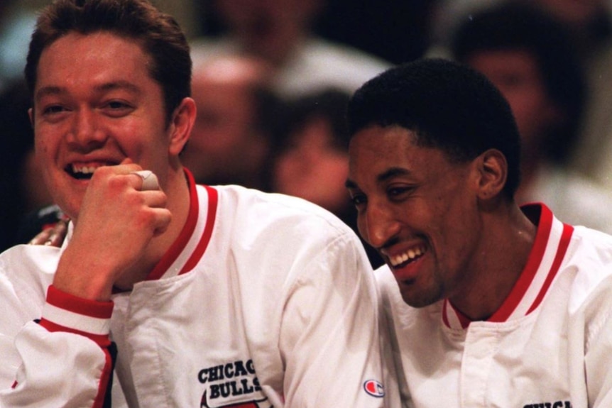 Chicago Bulls players Luc Longley and Scottie Pippen smile wide sitting courtside and wearing tracksuits