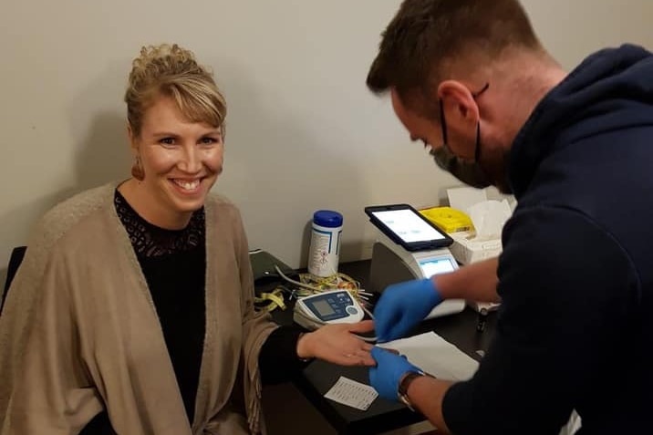 Woman sits in chair while man wearing gloves and mask collect fingerprick blood test.