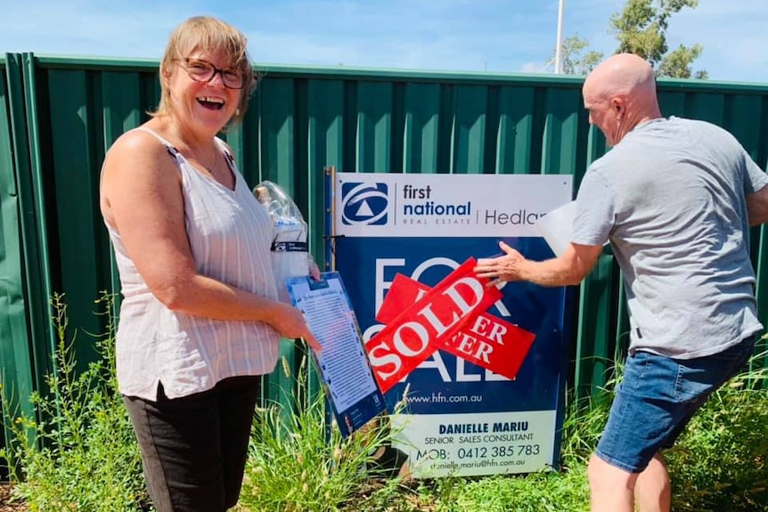 A woman and a man stand in front of a for sale sign on a green fence, with the man putting a red 'sold' sticker on the sign.