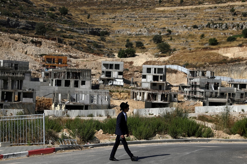 A young man in a black suit and black hat walks past half-constructed homes on an arid hill
