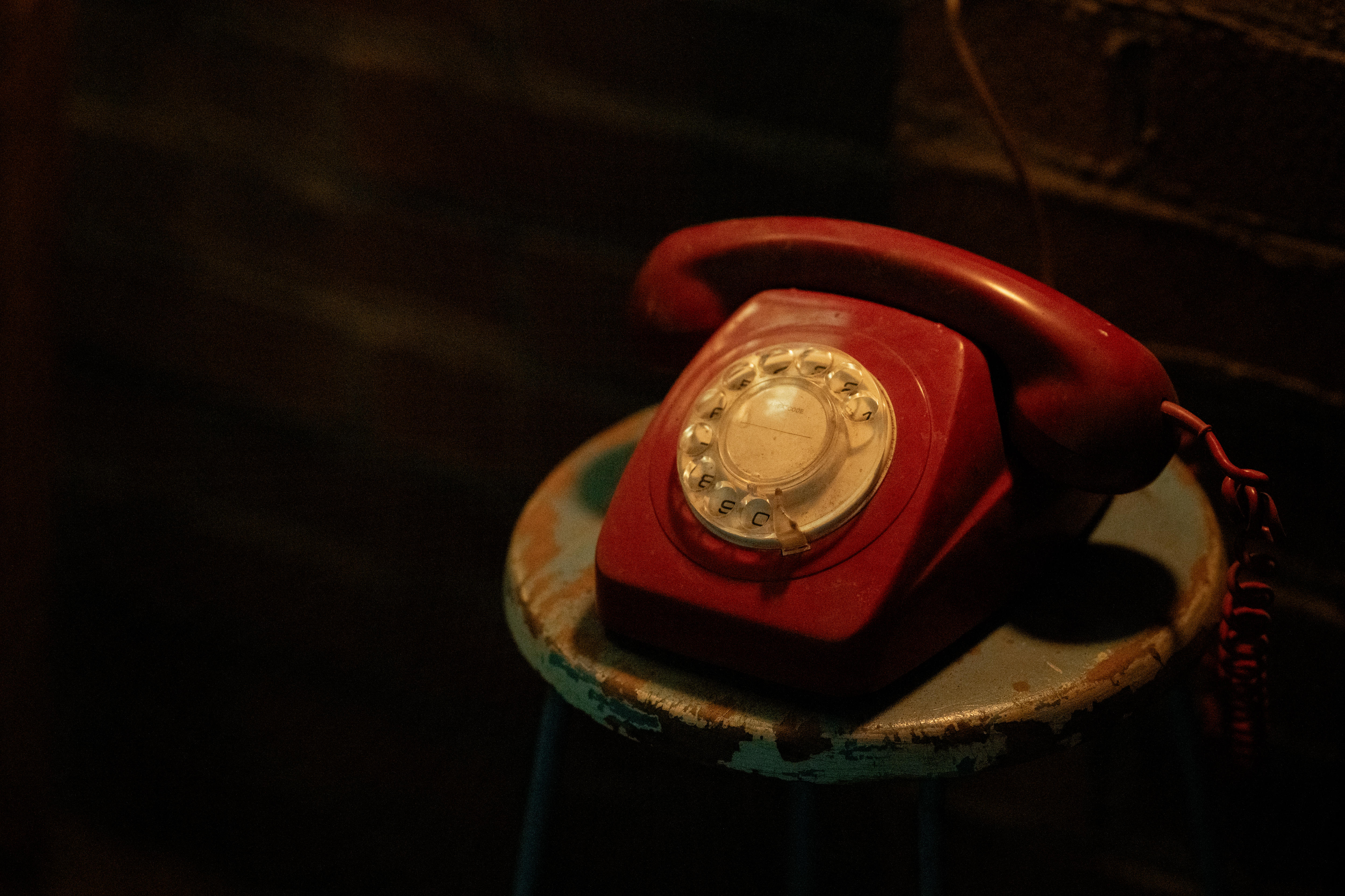 A retro red telephone is seen sitting on a chair in a dark room, with a light cast over the phone and nothing else.