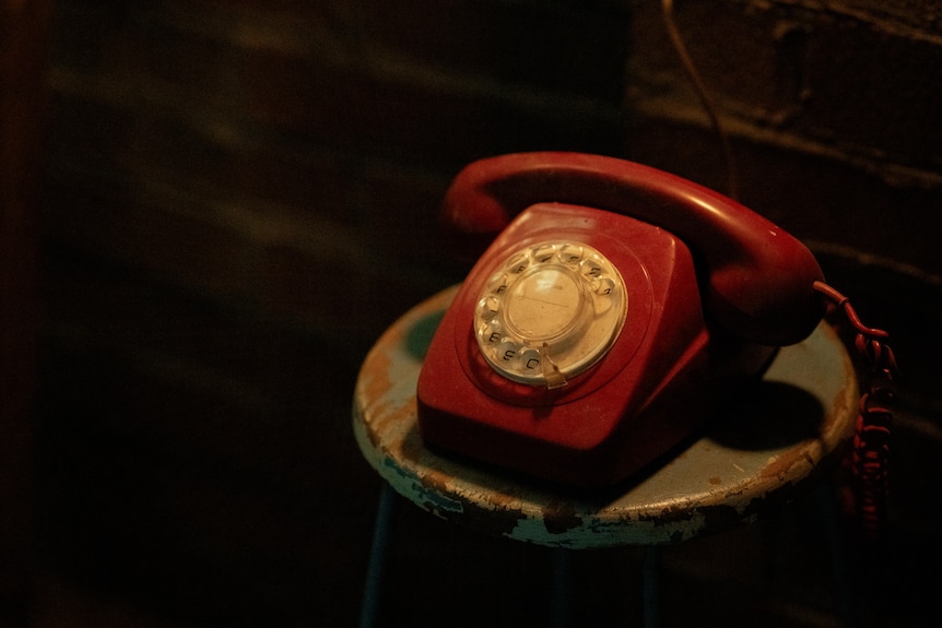 A retro red telephone is seen sitting on a chair in a dark room, with a light cast over the phone and nothing else.