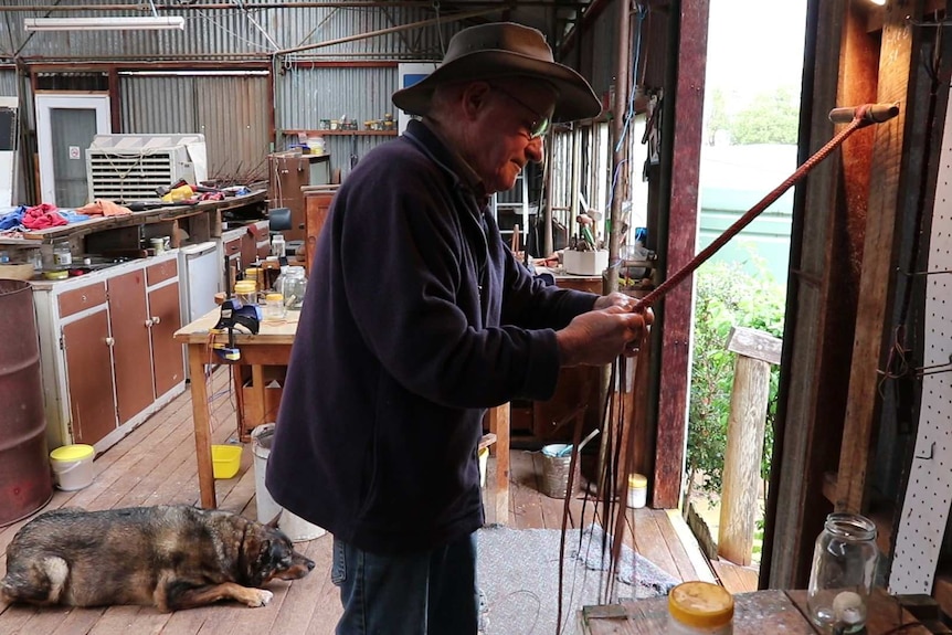 Dog on floor of shed on left, man standing weaving leather whip attached to hook on wall.