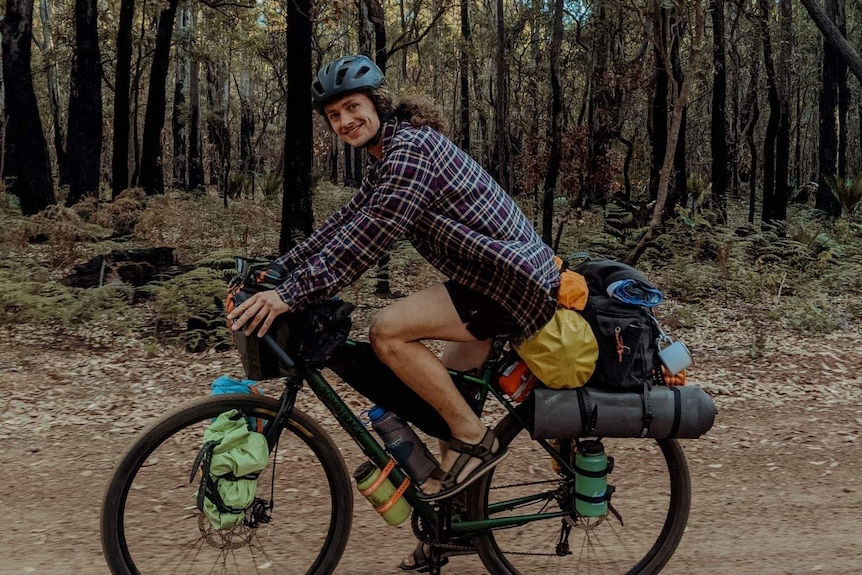 A man in a checkered shirt and blue helmet rides a bike loaded with camping equipment on a tree-lined, gravel road 
