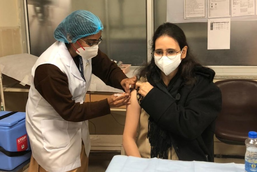 A woman with black hair and wearing a mask holds up her sleeve as another woman wearing scrubs administers a needle