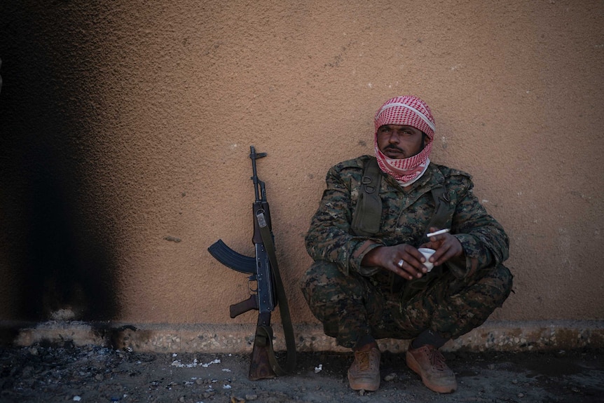 A man in a red checked scarf around his head and army gear crouches against a wall with gun next to him.