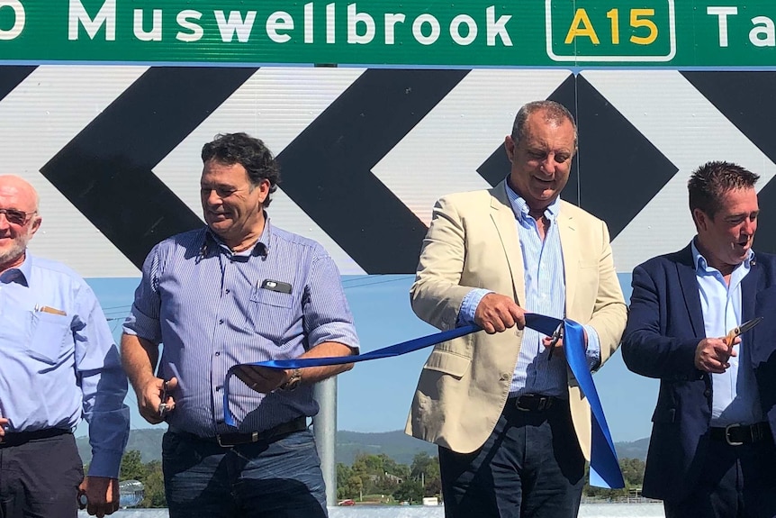 Three men standing in front of a road sign cutting a ribbon