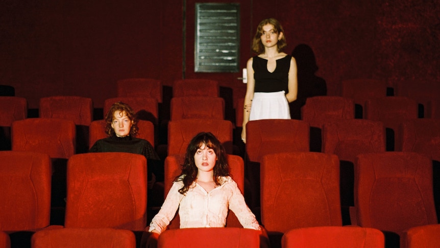 Three women sit in different rows in a theatre with red seats