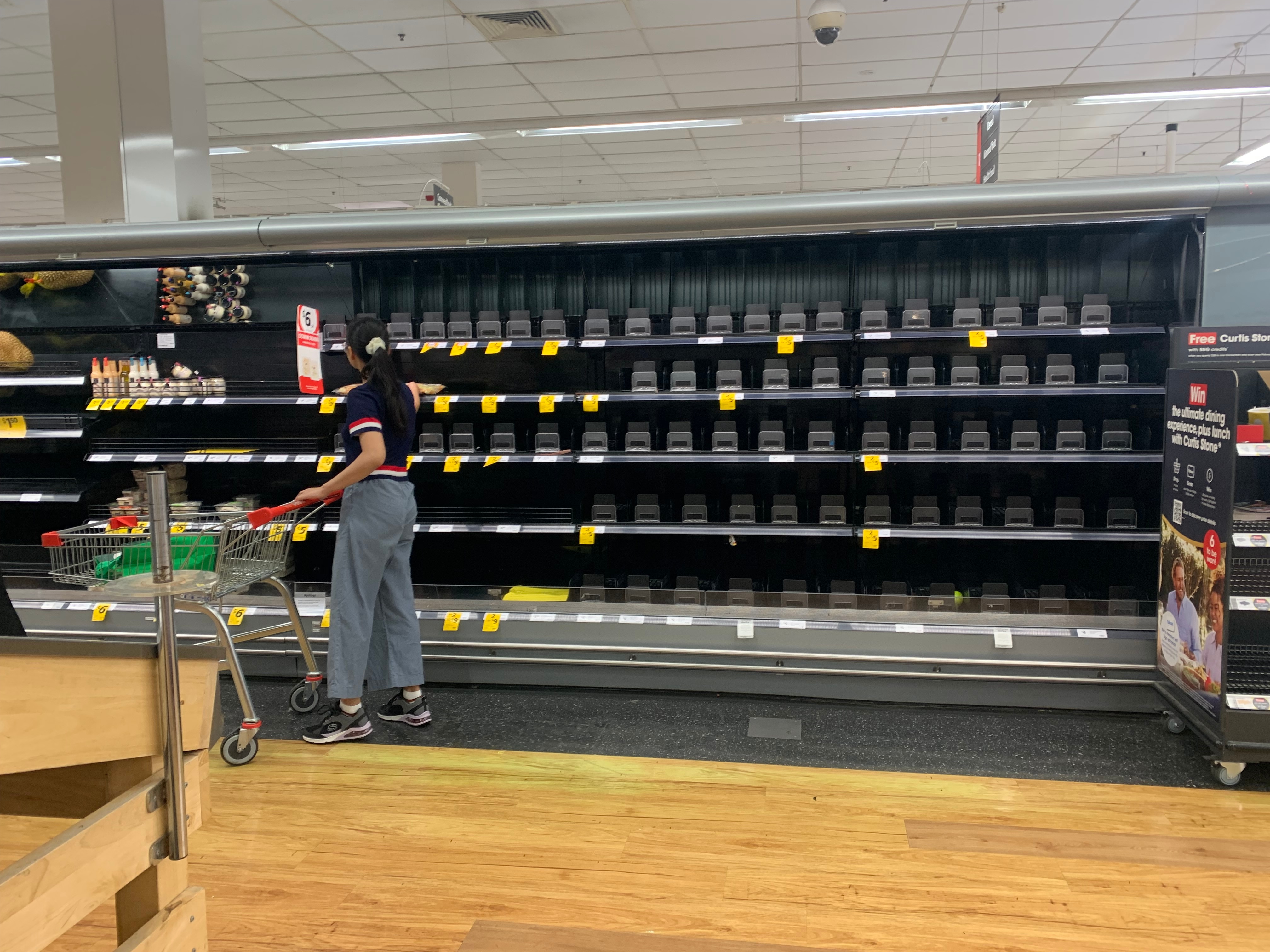 A lone shopper reaches for an item on an otherwise bare set of shelves in a supermarket's fresh produce section.