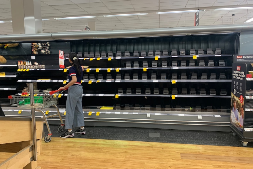 A lone shopper reaches for an item on an otherwise bare set of shelves in a supermarket's fresh produce section.