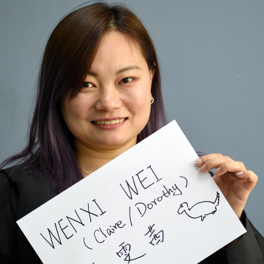 Woman faces camera holding sign reading "Wenxi Weir (Claire/Dorothy)