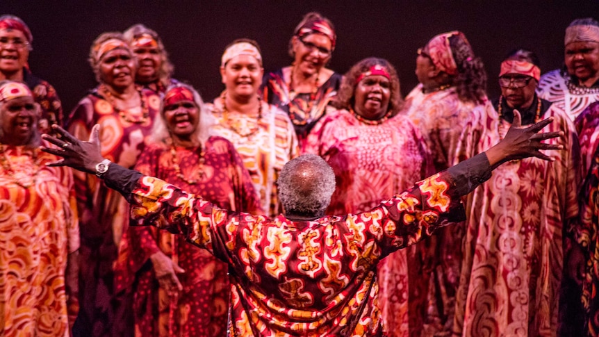 A man stands in front of a choir of women with his arms spread out