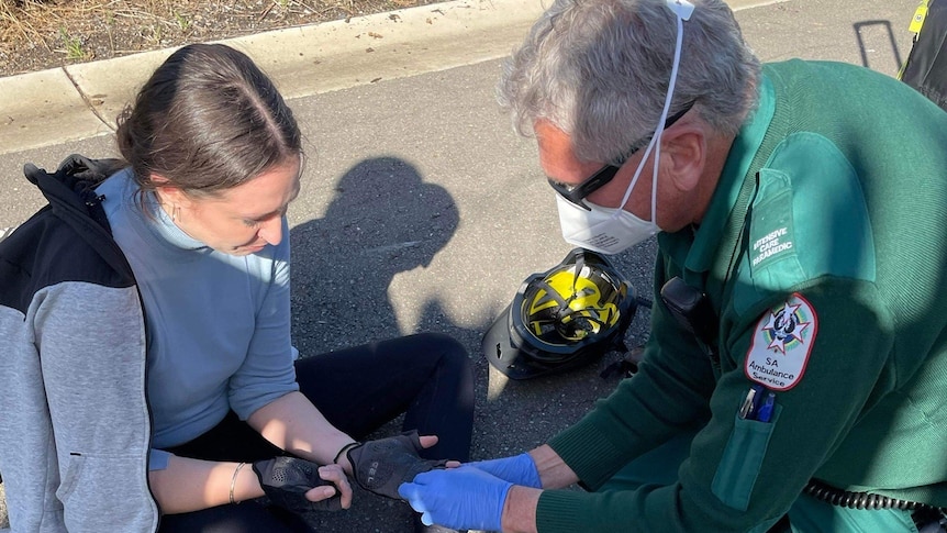 A woman sits on the floor while a man in green paramedic uniform and blue gloves inspects her hand