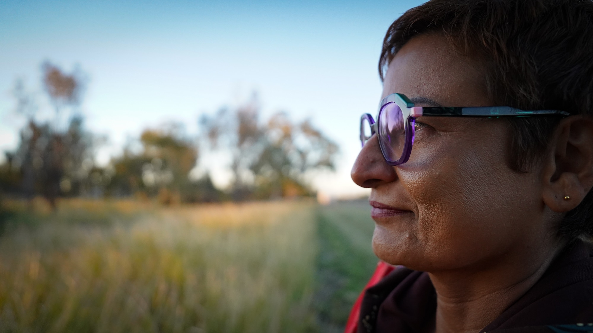 A close-up side profile of a woman wearing a neutral facial expression, looking over the vast outback landscape before her.