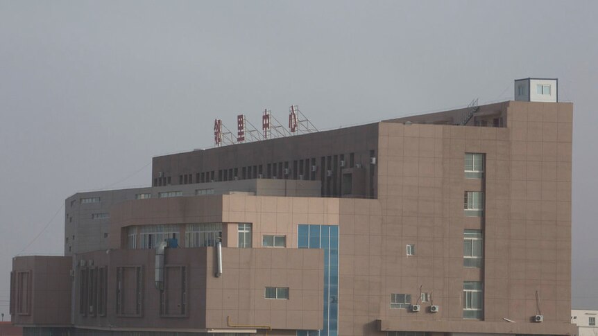 A large multi-storey brownish grey building behind a barbed wire fence with a large sign made up of red Chinese letters on top