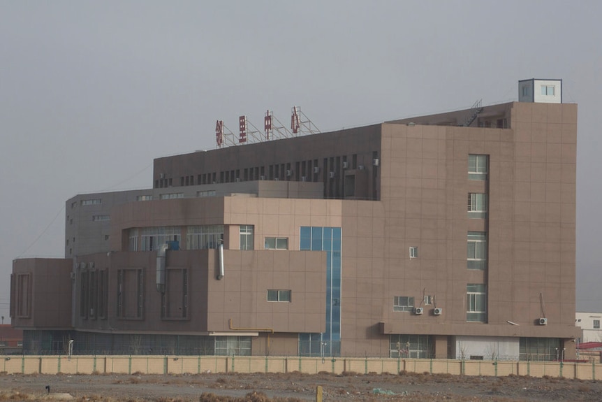 A large multi-storey brownish grey building behind a barbed wire fence with a large sign made up of red Chinese letters on top
