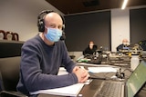 A man sits in a large room wearing a mask and a radio headset.