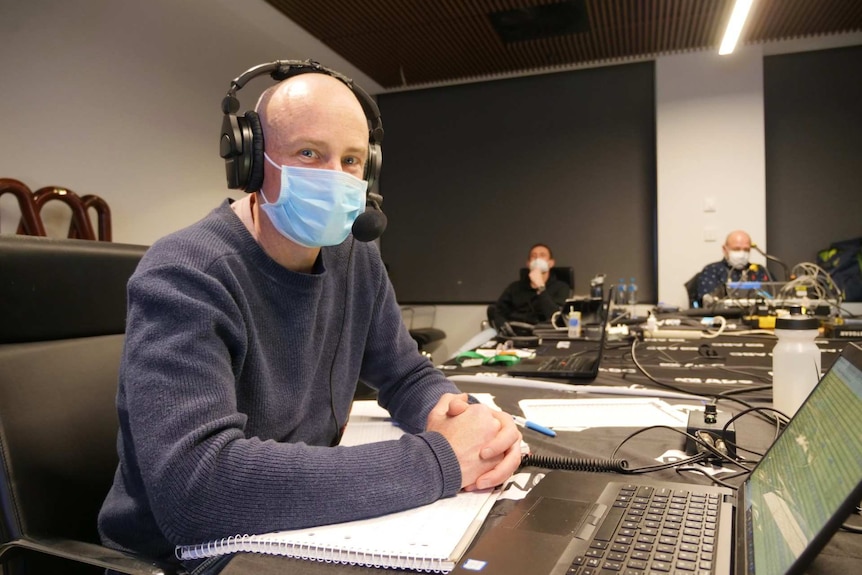 A man sits in a large room wearing a mask and a radio headset.