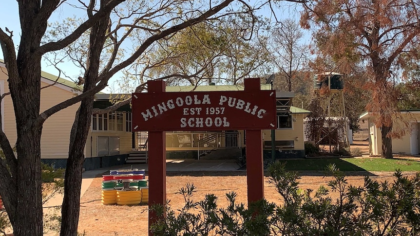 A sign saying Mingoola Public School Est 1957, under a tree in front of primary school buildings and a water tank