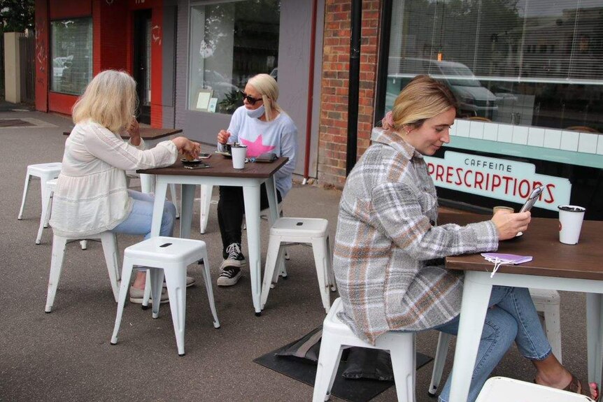 Three women sit at two different tables on a footpath outside a cafe, drinking coffee.