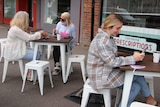 Three women sit at two different tables on a footpath outside a cafe, drinking coffee.