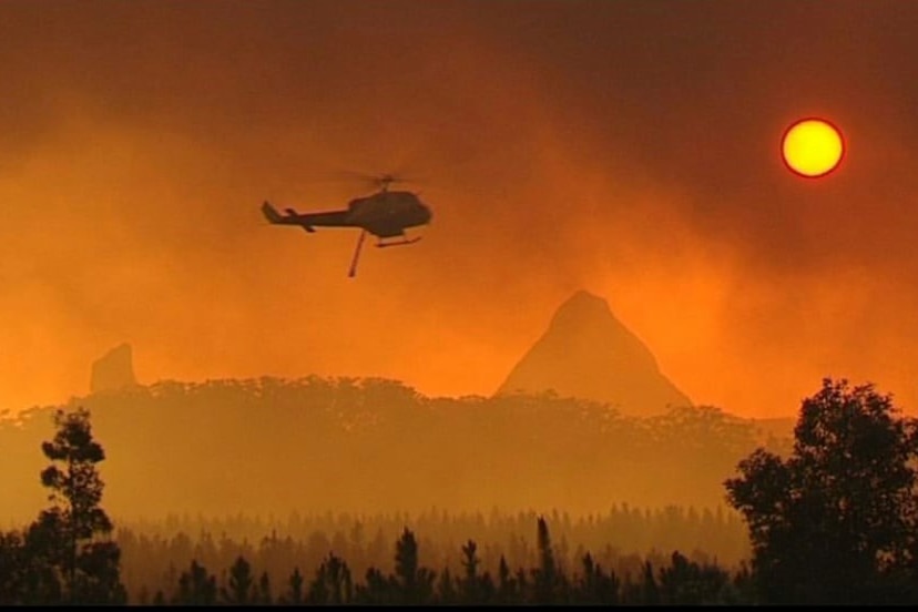 A waterbombing helicopter flies in an orange haze of smoke so thick that the sun looks like the moon.