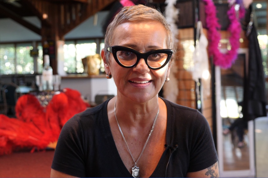 Woman in her fifties wearing cat eye classes, with cropped grey hair wearing a black T-shirt, feather boas behind her 