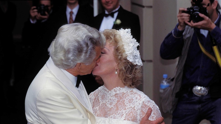 White haired man dressed in white suit kisses bride