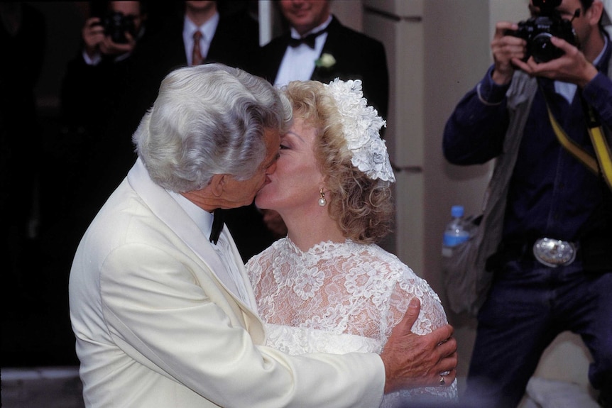White haired man dressed in white suit kisses bride