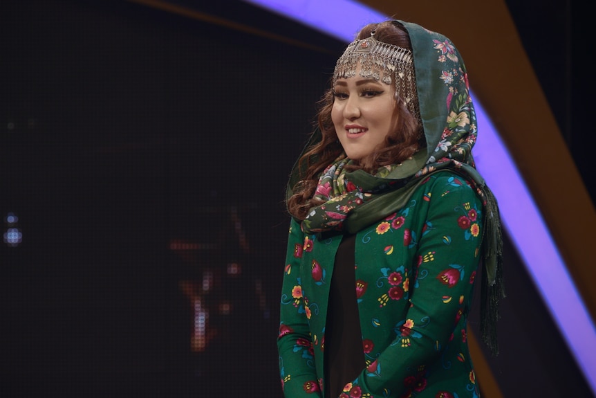 A young woman in traditional Afghan dress including a headscarf stands smiling on a studio stage