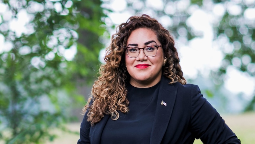 A woman with bright red lipstick, glasses and flowing dark hair smiling beneath a tree