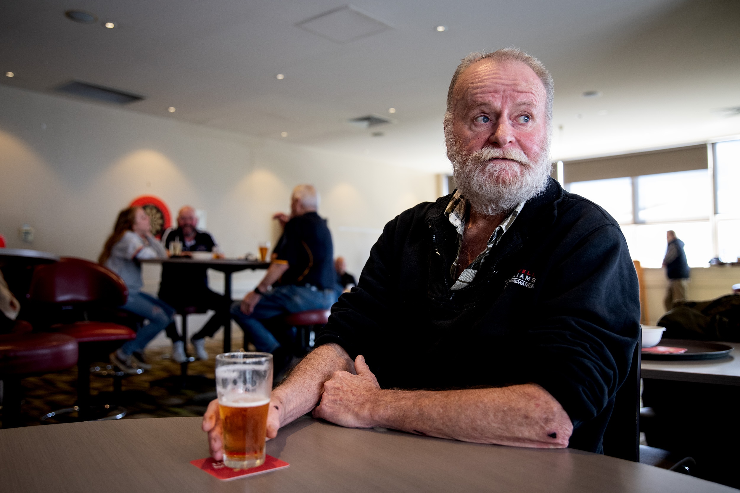 A man wearing a blue shirt with the sleeves rolled up sits at a table with his hand holding a glass of beer. People in the back