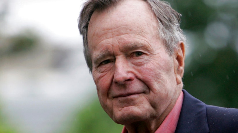 A man wearing a red shirt and navy jacket looks into the camera with blurry greenery behind him