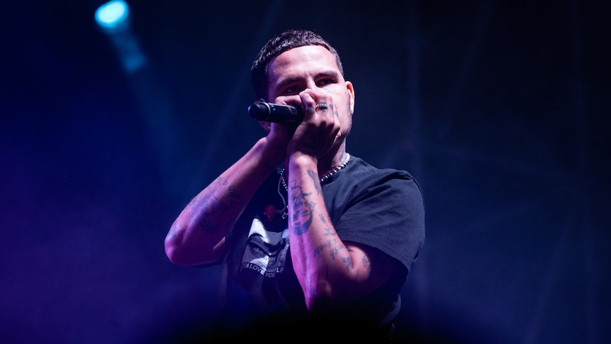 UK rapper Slowthai performing onstage in a black tshirt and holding a microphone to his mouth with both hands.