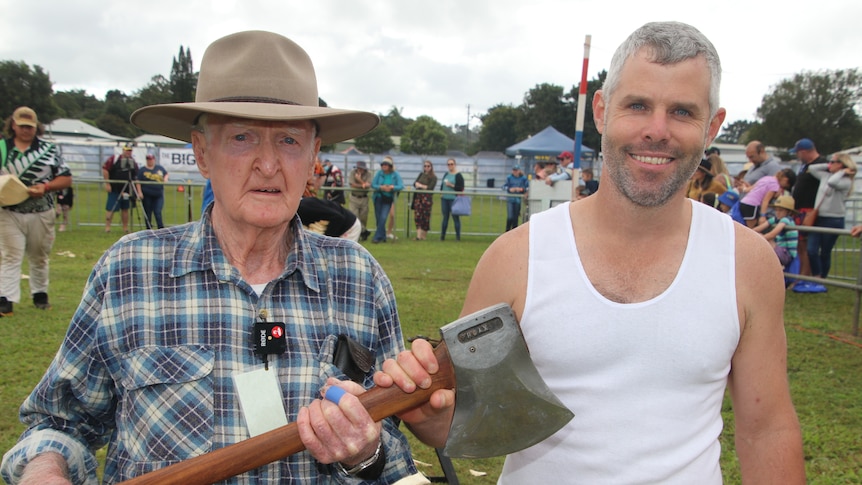 An elderly man in a broad-brimmed hat holds an axe as he stands beside a younger man who also has his hand on the axe