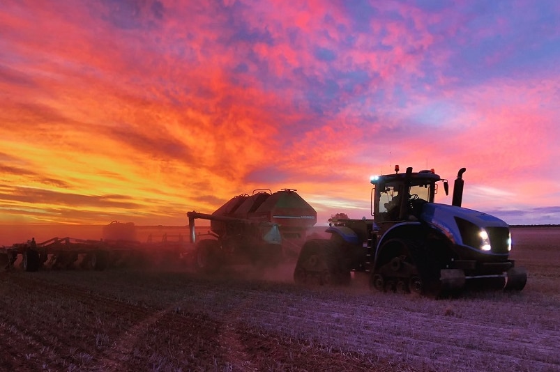 A tractor pulling crop planting equipment in a paddock sits underneath a vibrant and stunning sunset