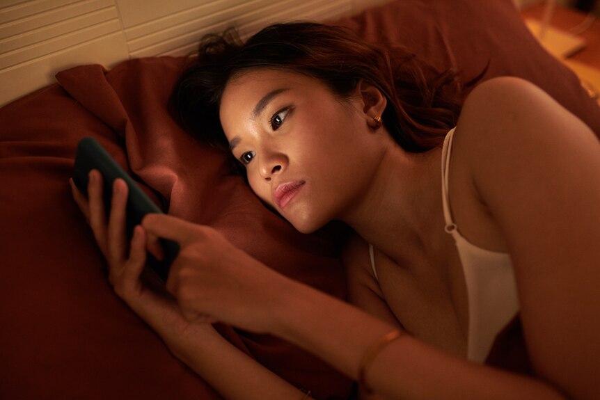A woman with long black hair lying down in bed looking at a smartphone