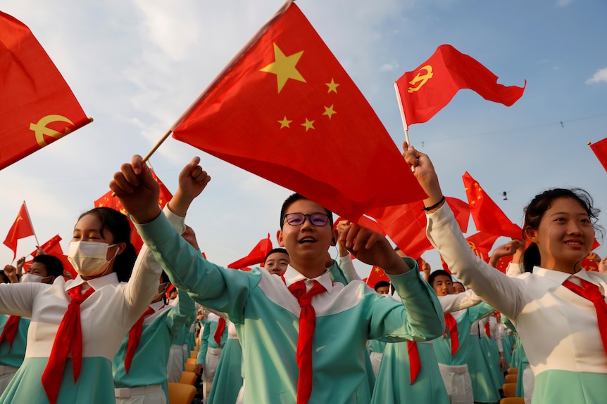 Primo piano di un folto gruppo di artisti in uniforme blu e bianca che sventolano le bandiere del Partito Comunista Cinese alle celebrazioni del centenario.  