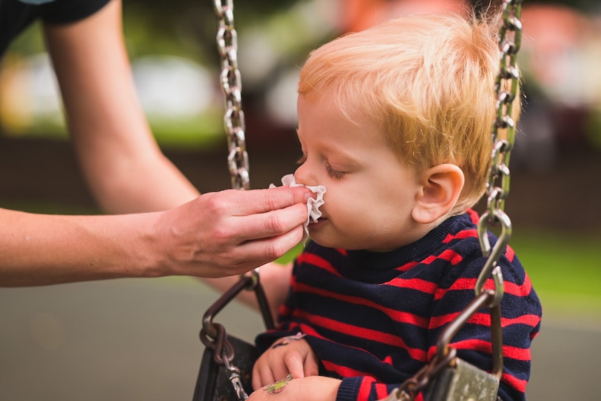 A young child sits on a swing while an adult holds a tissue to their nose to blow it 