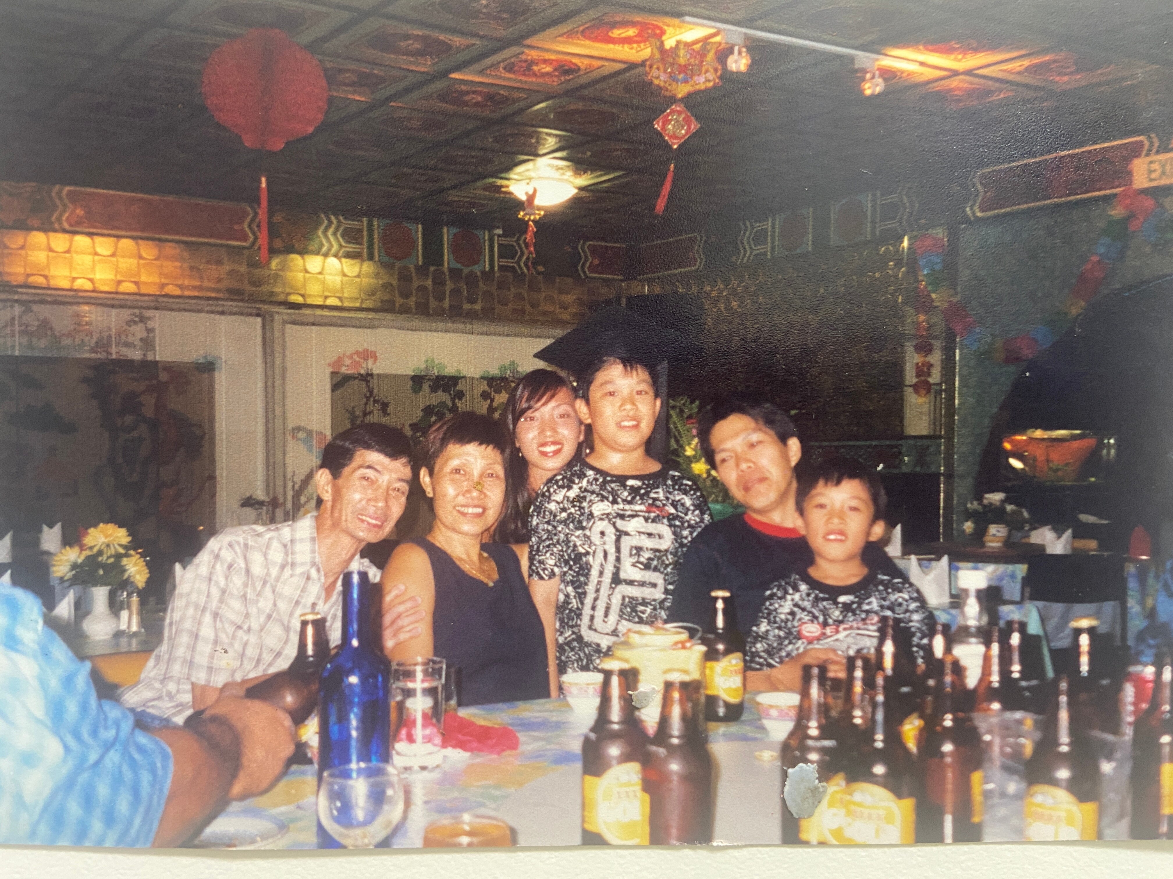 A young Chinese boy wearing a scholar's mortar board, posing for a photo with friends and family in a restaurant.